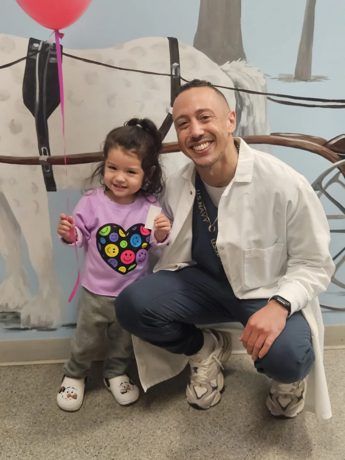 A Dental Care Kids dentist crouching beside a smiling toddler holding a balloon in the mural hallway
